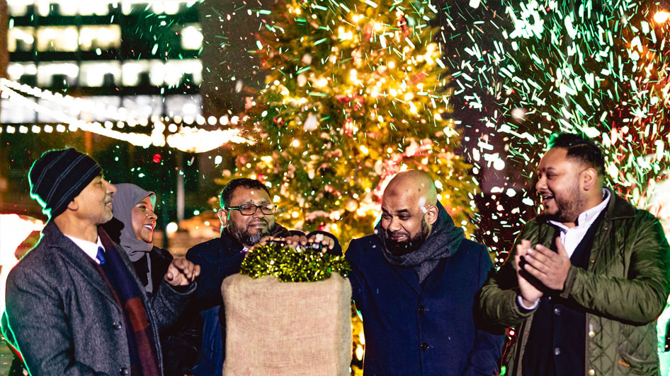 Group of people standing in front of a Christmas tree and snow, with a box and handle in front of them