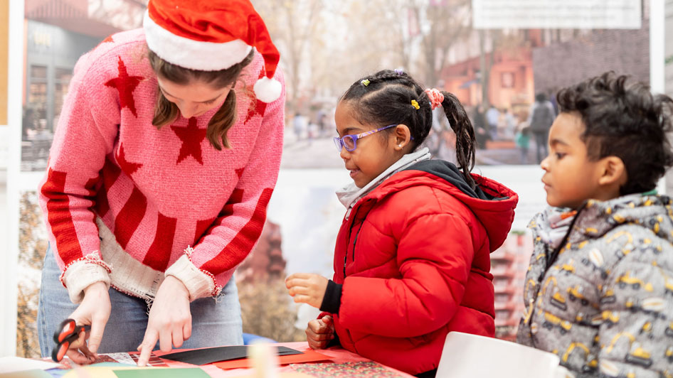 Woman wearing a santa hat demonstrating crafts to a young girl and boy